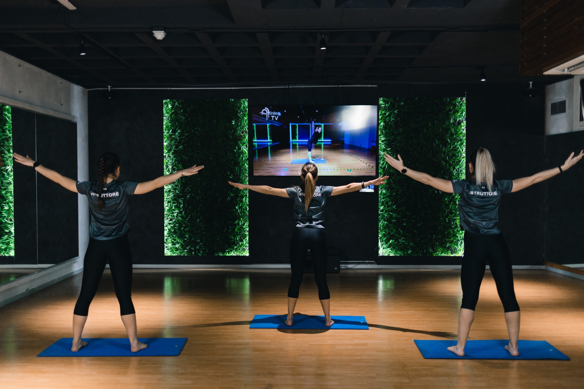 drei Frauen in schwarzer Kleidung im Fitnessstudio, die vor dem Fernseher trainieren