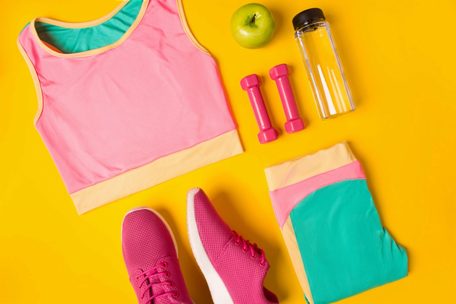 colorful gym merchandise including sports bra, leggings, weights water bottle and apple on yellow backgroud