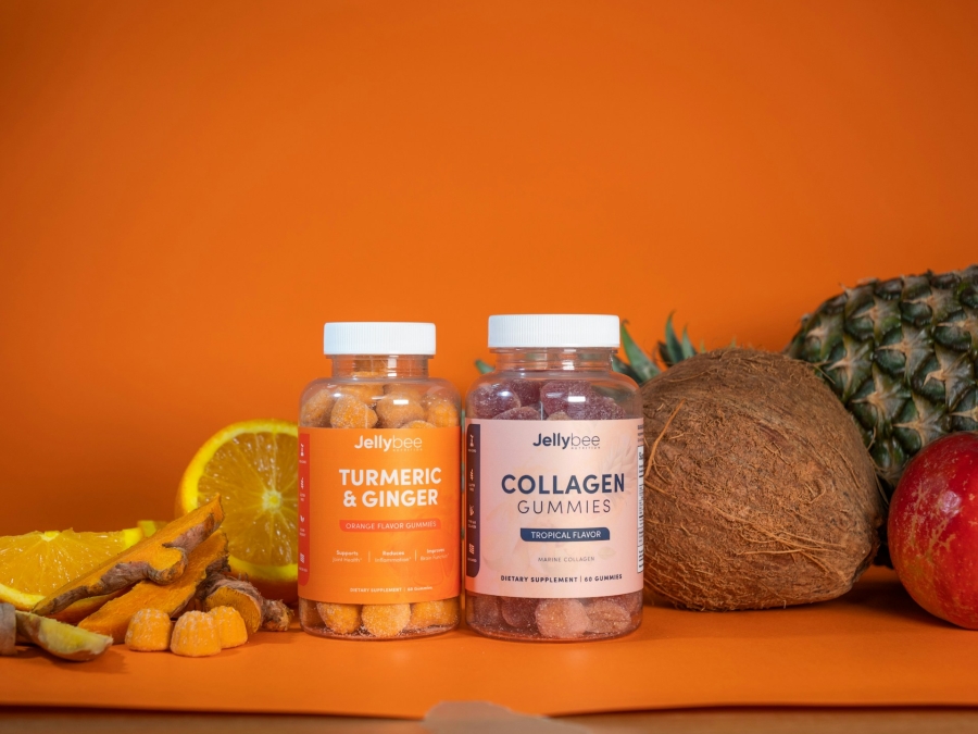 Transparent bottles of jelly supplements containing ginger, turmeric, and collagen, set against an orange background with fruits in the backdrop
